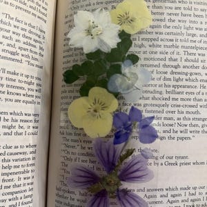 May include: A handmade bookmark featuring pressed flowers in white, yellow, and purple hues. The bookmark is placed in an open book, with the text visible. The flowers add a delicate, natural touch to the reading experience.