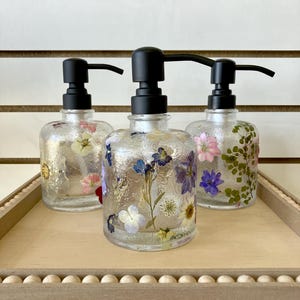 May include: Three clear glass soap dispensers with black pumps. Each dispenser is filled with pressed flowers and greenery. The dispensers are sitting on a wooden tray with a beaded edge.