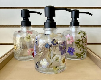 May include: Three clear glass soap dispensers with black pumps. Each dispenser is filled with pressed flowers and greenery. The dispensers are sitting on a wooden tray with a beaded edge.