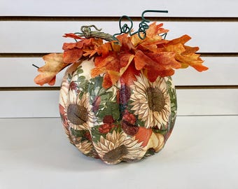 May include: A decorative pumpkin featuring a decoupage pattern of sunflowers, green foliage, and red berries. The top is adorned with a cluster of orange and yellow faux autumn leaves and a dark green wire curly vine stem.