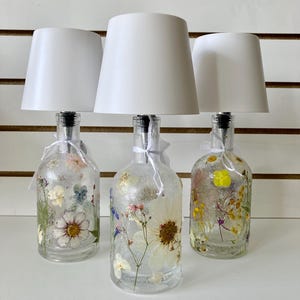 May include: Three decorative lamps made from clear glass bottles filled with dried flowers. Each bottle has a white lampshade and a silver-colored stopper. The lamps are arranged on a white surface.