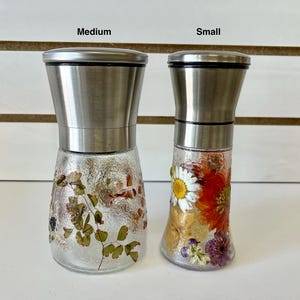 May include: Two glass spice grinders with stainless steel tops. One is medium-sized and decorated with dried leaves, the other is small and adorned with colorful dried flowers, including a daisy. Both have a textured glass base.