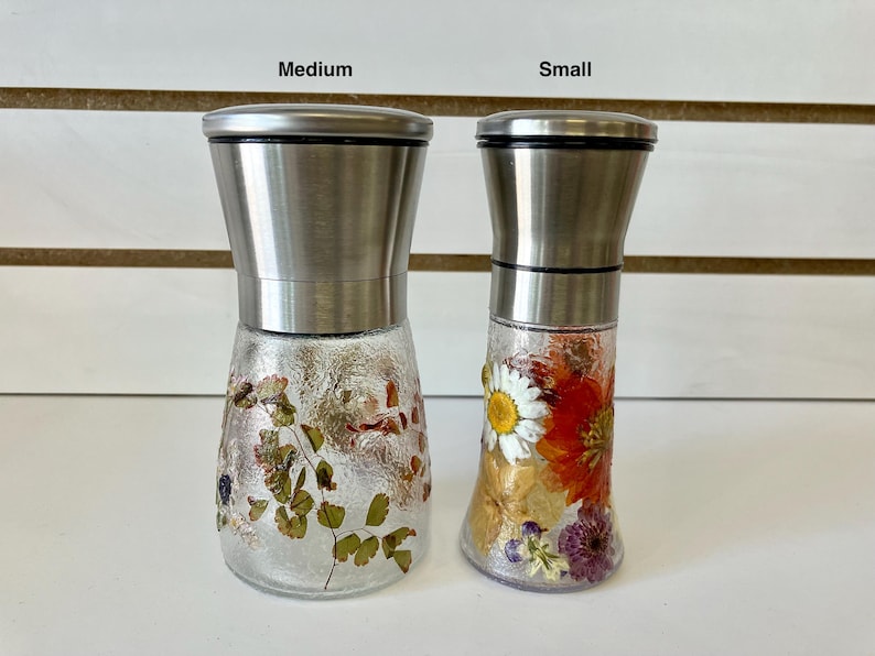 May include: Two glass spice grinders with stainless steel tops. One is medium-sized and decorated with dried leaves, the other is small and adorned with colorful dried flowers, including a daisy. Both have a textured glass base.