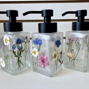 May include: Three clear glass soap dispensers with black pumps. Each dispenser is decorated with pressed flowers in shades of white, blue, and pink. The dispensers are square-shaped.