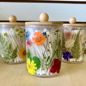May include: Three clear glass jars with wooden lids filled with cotton swabs and pressed flowers. The jars are arranged in a row on a wooden surface. The flowers are a variety of colors, including pink, purple, yellow, and orange.