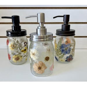 May include: Three clear glass mason jar soap dispensers with pressed flowers inside. The dispensers have black or silver pump tops. The flowers are in shades of yellow, red, blue, and white.