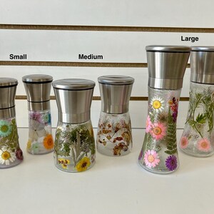 May include: A collection of glass spice grinders with stainless steel tops, each filled with dried flowers. The grinders are labeled "Small", "Medium", and "Large". The flowers are various colors, including pink, yellow, and white.