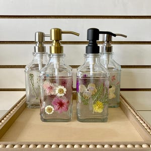 May include: Four glass soap dispensers with different colored pumps. Each dispenser is filled with clear liquid and dried flowers. The dispensers are sitting on a wooden tray with a beaded edge.