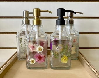 May include: Four glass soap dispensers with different colored pumps. Each dispenser is filled with clear liquid and dried flowers. The dispensers are sitting on a wooden tray with a beaded edge.