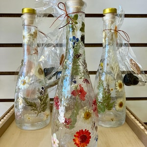 May include: Three clear glass bottles with cork stoppers and gold lids. Each bottle is filled with pressed flowers and greenery. The bottles are arranged in a row on a wooden surface.