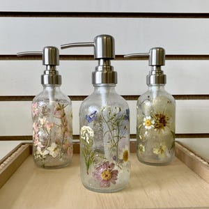 May include: Three clear glass soap dispensers with stainless steel pumps. Each dispenser is filled with pressed flowers and has a different floral design.
