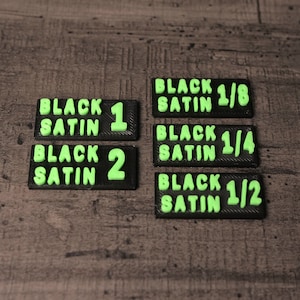 Peut inclure: Six étiquettes rectangulaires noires avec du texte vert. Les étiquettes indiquent "Black Satin 1", "Black Satin 2", "Black Satin 1/8", "Black Satin 1/4" et "Black Satin 1/2".