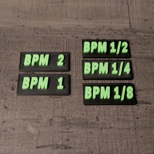 Könnte beinhalten: Eine Ansammlung schwarzer rechteckiger Etiketten mit erhabenem, neongrünem Text. Die Etiketten zeigen "BPM 2", "BPM 1", "BPM 1/2", "BPM 1/4" und "BPM 1/8". Sie liegen auf einer strukturierten, grauen Oberfläche.