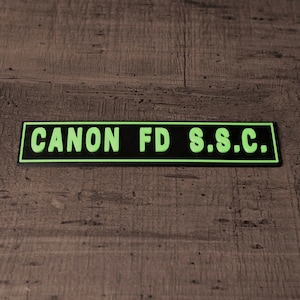 Może przedstawiać: Prostokątna czarna naszywka z napisem "CANON FD S.S.C." w jaskrawozielonych literach. Naszywka ma czarną obwódkę i jest umieszczona na tle usłojenia drewna.