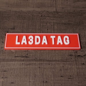 Peut inclure: Une plaque rectangulaire rouge avec une inscription blanche qui dit "LA3DA TAG".