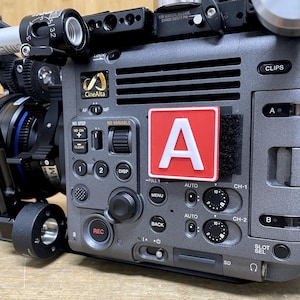 Peut inclure: Gros plan d'une caméra vidéo professionnelle avec une étiquette rouge "A" fixée sur le côté. La caméra possède une variété de boutons et de molettes, notamment un bouton d'enregistrement, un bouton de menu et un écran d'affichage. La caméra est également équipée d'un objectif fixé à l'avant.