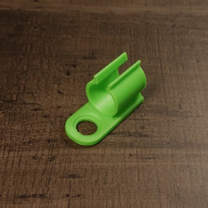 Puede incluir: Un clip de cable de plástico verde brillante con un agujero redondo y una sección curva para sujetar cables.