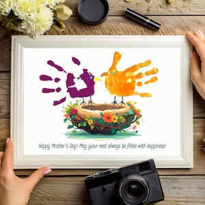 Puede incluir: Una impresión enmarcada con un diseño de arte de huellas de manos de dos pájaros en un nido. Los pájaros son morados y naranjas y el nido está decorado con flores. El texto "¡Feliz Día de la Madre! ¡Que tu nido siempre esté lleno de felicidad!" está impreso debajo de la imagen.