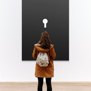 May include: A person stands in front of a black canvas with a white keyhole shape in the center. The person is wearing a brown jacket, black pants, and a white drawstring bag.