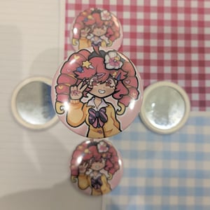 May include: Pink and yellow cartoon character buttons with a peace sign gesture. The character has pink hair and is wearing a yellow jacket. Two small round mirrors are also in the image. The background includes a red and white checkered pattern and a blue and white checkered pattern.