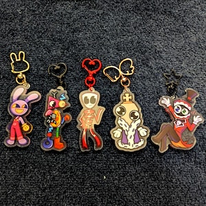May include: Five acrylic keychains featuring cartoon characters with unique designs. The first is a purple rabbit, the second a colourful geometric robot, the third a red abstract figure, the fourth a king in robes, and the fifth a ringmaster.