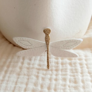 May include: A white felt dragonfly with a gold glitter body. The dragonfly is a decorative accent for a home or craft project.