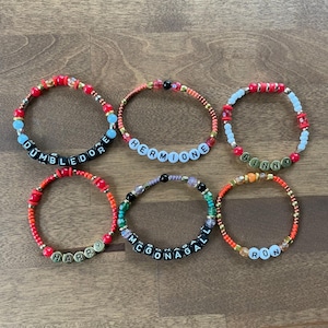 May include: Six colorful beaded bracelets with different names spelled out in black letter beads. The names are Dumbledore, Hermione, Ginny, Harry, McGonagall, and Ron.