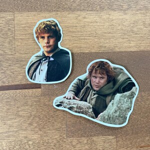 May include: Two stickers featuring characters from The Lord of the Rings. The first sticker shows a young man with brown hair and a brown cloak. The second sticker shows a man with brown hair and a brown beard, wearing a brown cloak and holding onto a rock.