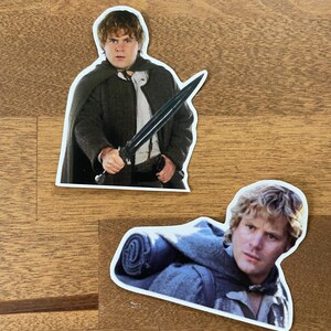 May include: Two stickers featuring characters from the Lord of the Rings movie franchise. The first sticker shows a character holding a sword, while the second sticker shows a character with a backpack.