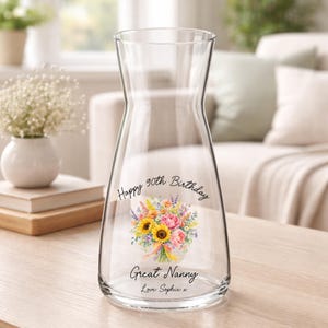May include: A clear glass carafe featuring a printed watercolour bouquet of sunflowers and pink flowers. The text reads Happy 90th Birthday, Great Nanny, and Love Sophie x. The vessel has a wide base and a tapered neck with a flared rim.