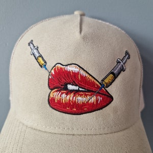 May include: A beige trucker hat with a red embroidered pair of lips with two yellow and blue syringes piercing through them.