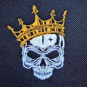 Puede incluir: Una calavera bordada en blanco que lleva una corona dorada. La corona tiene un diseño de flor de lis en la parte superior y la palabra "RIP" está bordada en la banda.