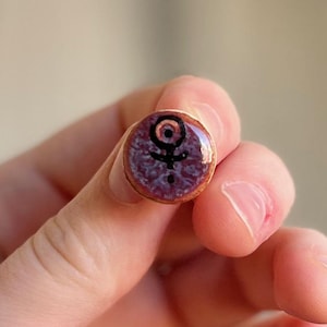 Op de afbeelding: Een kleine, ronde, koperkleurige pin met een paarse achtergrond en een zwart Venus-symbool. De pin wordt in de hand van een persoon gehouden, met een vage achtergrond.