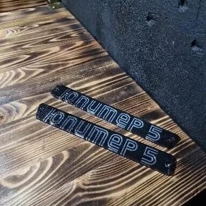 May include: Two black metal plates with the text "nummer 5" in white lettering. The plates are on a wooden surface.