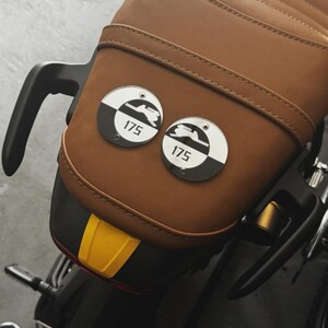 May include: A brown leather motorcycle seat with two circular metal badges. Each badge has a black and white design with the number "175" and a motorcycle silhouette. The seat has a quilted pattern.