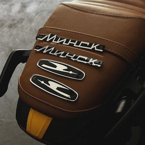 May include: Close-up of a brown motorcycle seat with the word "Минск" in silver lettering. Below are two silver and black emblems. The seat has a quilted pattern and a yellow and black accent.