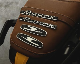 May include: A close-up of a brown motorcycle seat featuring silver "Минск" lettering and two silver and black emblems. The seat has a quilted design and a yellow and black accent.