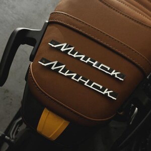 May include: Close-up of a motorcycle seat with a brown leather finish and quilted stitching. Two silver and black "Минск" emblems are affixed to the seat. The seat has a black handle and a yellow accent.