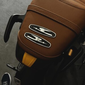 May include: Close-up of a brown leather motorcycle seat with a quilted pattern. Two silver and black emblems are affixed to the seat. A portion of the motorcycle's black frame and a yellow accent are visible.