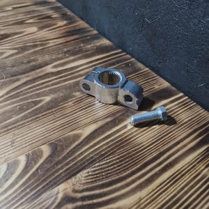 May include: A silver-colored metal component with a circular center and two holes, accompanied by a silver bolt. The parts are displayed on a wooden surface with a dark background.