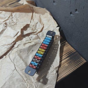 Puede incluir: Un portafusibles negro con una variedad de fusibles de colores. Los fusibles son de varios colores, incluyendo azul, amarillo, rojo y naranja. El soporte está sobre una superficie de papel marrón arrugado.