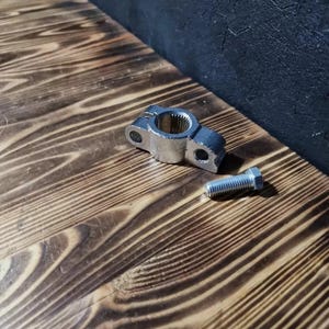 Op de afbeelding: Een zilveren metalen pijpklem met een schroef. De klem bevindt zich op een houten oppervlak.