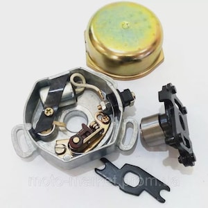Puede incluir: Un conjunto de piezas del sistema de encendido de motocicletas, que incluye una carcasa de distribuidor plateada con componentes internos, una tapa dorada y un rotor negro. Las piezas están dispuestas sobre una superficie blanca.