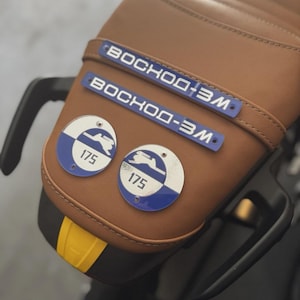 May include: A brown motorcycle seat with two blue and white circular emblems and two blue rectangular emblems. The circular emblems have the number 175. The rectangular emblems have the text "BOCHOO-3M".