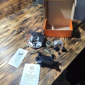 May include: A collection of car parts, including a black and silver ignition system component, metal brackets, and a small orange box. Two instruction manuals are also present, along with other small parts. The items are arranged on a wooden surface.