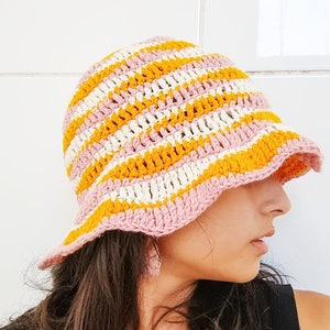 May include: A crocheted bucket hat with pink, white and yellow stripes. The hat is being worn by a person with long brown hair.