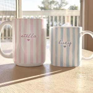 May include: Two ceramic mugs with white handles sit on a wooden table. One mug is pink and white striped with the name "stella" and a heart. The other is blue and white striped with the name "lucy" and a heart.