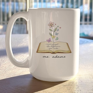 May include: White ceramic mug featuring a floral and butterfly design above an open book illustration. The text on the book reads "never forget the difference you make" and "ms. adams" is written below. The mug has a large handle.