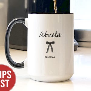 May include: A white ceramic mug with a black handle and rim is being filled with coffee. The mug has the word "Abuela" and a bow graphic printed on it, with "est. 2026" below. A coffee maker and kettle are in the background.