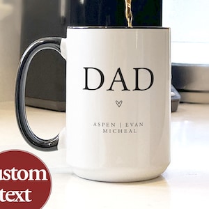 May include: White ceramic mug with black rim and handle, featuring the word "DAD" in black, a small heart, and the names "Aspen | Evan Micheal". Coffee is being poured into the mug.